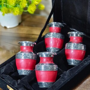 May include: Five small red urns with silver bands and lids, displayed in a black velvet box. The urns are vase-shaped and designed for keepsakes. Each urn is approximately 7 cm tall.