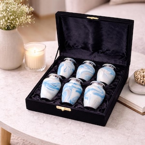 May include: Six small urns with a blue and white swirl pattern, displayed in a black velvet box. Each urn has a silver-coloured rim. The box is on a round, light-coloured table. The scene is softly lit.