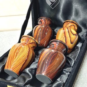 May include: Four wooden urns with a glossy finish, presented in a black velvet box. The urns feature a warm, reddish-brown wood grain pattern. The box has a gold clasp and a logo on the inside.