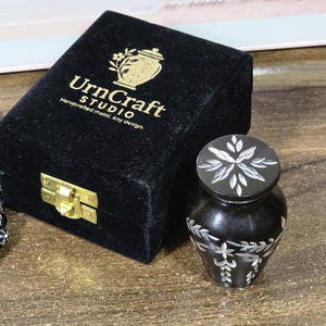 May include: A black urn with white floral patterns and a matching lid, beside a black velvet box with gold fittings. The box is labelled "UrnCraft STUDIO" and "Handcrafted metal. Any design."