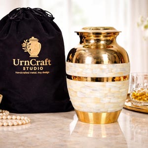 Könnte beinhalten: Eine gold-weiß Urne mit einer schwarzen Tasche. Die Urne hat eine polierte Goldoberfläche und eine weiße, perlmuttartige Einlage. Auf der schwarzen Tasche steht "UrnCraft STUDIO".