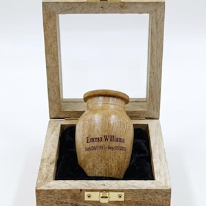 May include: A wooden urn with a glass lid, displayed in an open wooden box. The urn is light brown with a rounded shape and a wooden lid. The urn is engraved with the name "Emma Williams" and dates Feb/26/1951-Sep/05/2023.