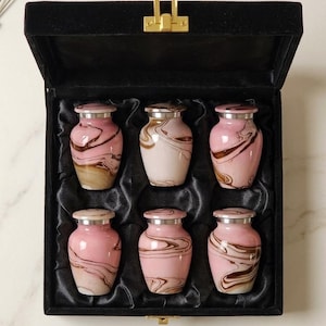 May include: A set of six small urns in a black velvet box. The urns are pink with brown marbling and silver rims. The box has a gold clasp. A sprig of lavender and a white petal are in the background. The urns are for keepsakes.