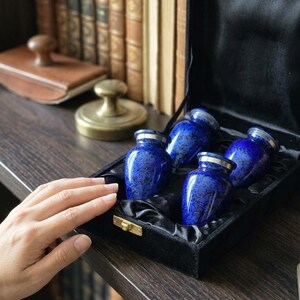 May include: A set of four small, cobalt blue cremation urns with mottled black patterns and silver-toned rims, presented inside a black velvet-lined display box with a gold-coloured metal latch closure.