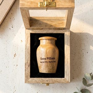 May include: A wooden urn with a glass-topped box. The urn is cylindrical with a rounded top and is engraved with the name "Emma Williams" and dates "Feb/26/1951- Sep/05/2023". The box has a brass clasp and is lined with black velvet.