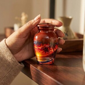 May include: A small, cylindrical metal urn featuring a vibrant sunset scene with silhouettes of palm trees, a bright yellow sun, and deep orange, red, and purple clouds over a calm ocean. The urn has a smooth, glossy finish and a matching lid.