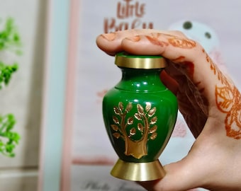 Puede incluir: Una pequeña urna verde con un diseño de árbol dorado. La urna tiene una tapa y una base doradas. Se sostiene en una mano con tatuajes de henna. Una caja negra con un emblema dorado está en primer plano.
