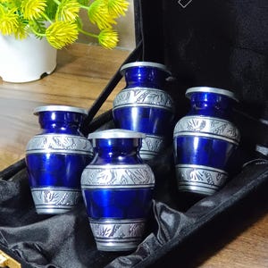 May include: Four small, blue urns with silver decorative bands, presented in a black velvet box. The urns have a metallic finish and a classic design. Ideal for memorial keepsakes, they are a thoughtful tribute.