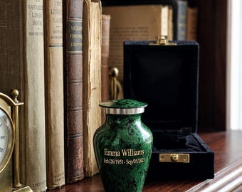 Puede incluir: Una pequeña urna funeraria verde con patrón de mármol y tapa plateada. La urna está grabada con el texto Emma Williams y las fechas Feb/26/1951-Sep/05/2023. Se encuentra junto a una caja negra forrada de terciopelo sobre una superficie de madera.