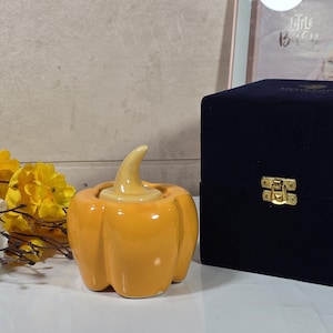 May include: A ceramic pumpkin-shaped candle holder in shades of orange and yellow. The candle holder has a stem and a round opening. A black velvet box with a gold clasp is next to the candle holder.