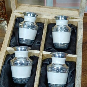 May include: A wooden box with a glass lid contains four white and silver decorative urns. Each urn has a silver lid and base with ornate detailing. The urns are displayed on black fabric.
