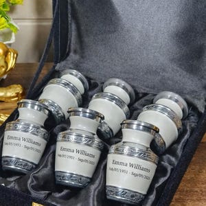 May include: A set of six small, white and silver urns, each inscribed with "Emma Williams" and dates. The urns are displayed in a black velvet-lined box with a gold clasp. The urns have a decorative silver band.