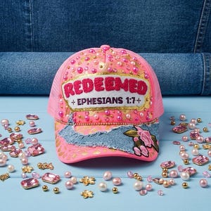 Puede incluir: Gorra rosa con la palabra "REDEEMED" y "Ephesians 1:7+" bordada en la parte delantera. La gorra está decorada con perlas y pedrería. Hay más perlas y pedrería esparcidas alrededor de la gorra.
