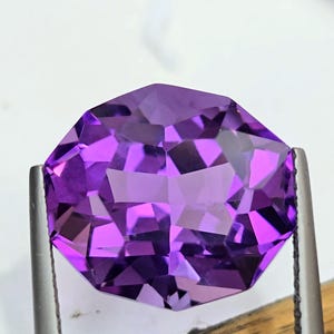 May include: A faceted, octagonal amethyst gemstone held by metal tweezers. The gem is a deep purple color with multiple facets that reflect light. The background is white.