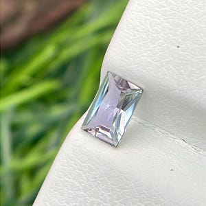 May include: A rectangular, faceted gemstone with a pale lavender hue. The gem is set against a white, textured surface, with green foliage in the background. The gem's facets reflect light, creating a sparkling effect.