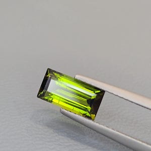 May include: A rectangular, baguette-cut gemstone with a vibrant, translucent lime-green color. The stone features sharp, clean facets that reflect light, creating a bright, polished appearance. It is held by a pair of silver-toned metal tweezers.