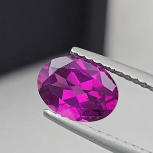 May include: A faceted, oval-shaped gemstone held by tweezers. The gem is a vibrant magenta color, with multiple facets catching the light. The background is blurred, with a dark shape on the left and a light surface.