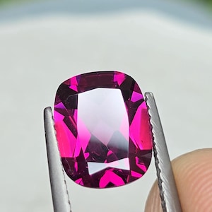 May include: A cushion-cut, faceted gemstone in shades of magenta and pink is held by a pair of tweezers. The gem has a high degree of clarity and brilliance, reflecting light. The background is blurred, suggesting a natural setting.