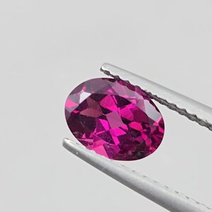 May include: A vibrant, oval-cut gemstone in a deep magenta hue is held by silver tweezers against a white background. The gemstone's facets reflect light, creating a sparkling effect. This image showcases a precious stone, likely for jewelry.