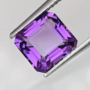 Op de afbeelding: Een gefacetteerde, vierkant geslepen amethist edelsteen, vastgehouden door een zilveren pincet. De edelsteen heeft een dieppaarse kleur met een lichter centrum, wat de helderheid en slijpvorm laat zien. De pincet staat schuin tegen een witte achtergrond.