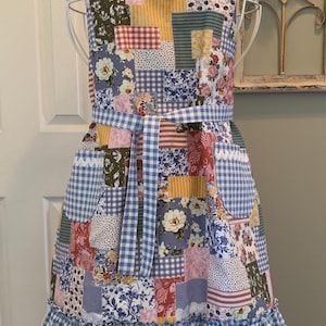 May include: A patchwork apron with a blue and white gingham trim and a ruffled bottom. The apron has a variety of floral and geometric patterns in shades of blue, pink, yellow, and green.