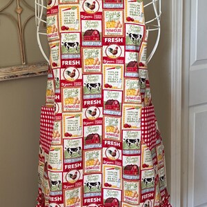 May include: A kitchen apron featuring a repeating grid pattern of farm-themed illustrations including cows, roosters, barns, and text like "Farmhouse Organics" and "Fresh." The apron has red and white gingham straps, pockets, and a ruffled hem.