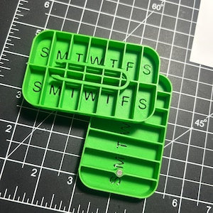 May include: Bright green weekly pill organizer with two sections, each labeled with the days of the week: S, M, T, W, T, F, S. The organizer is made of plastic and has a snap closure. The background is a black cutting mat with grid lines.