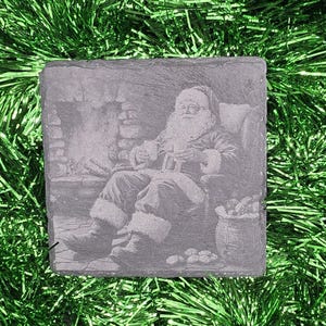 May include: A square, gray slate coaster featuring an etched image of Santa Claus relaxing in a chair by a fireplace. Santa is holding a mug and there is a sack of gifts nearby. The coaster is surrounded by green tinsel.