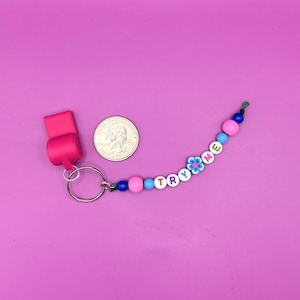 May include: A pink whistle attached to a silver metal keyring and a string of colorful beads. The beads include white letter beads spelling TRY ME, a blue flower-shaped bead, and various round beads. A US quarter coin is shown for scale.