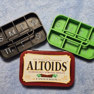 May include: Two 3D-printed pill organisers, one black and one green, with the days of the week marked. An Altoids cinnamon mint tin is also present. The tin is red with gold lettering and a cinnamon stick graphic.