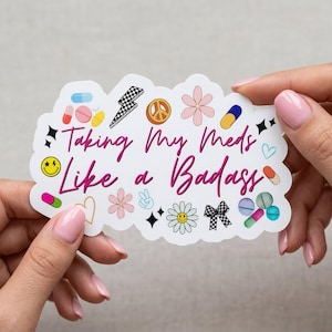 May include: A white sticker with the text "Taking My Meds Like a Badass" in pink script. The sticker is decorated with various colorful illustrations, including pills, flowers, and a peace sign.