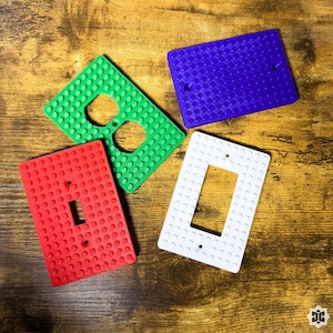 May include: Four rectangular wall plates with a raised stud texture resembling building blocks. The set includes one green duplex outlet cover, one solid purple blank cover, one red toggle switch cover, and one white rocker switch cover.