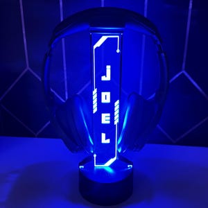 May include: A black headset stand with blue LED lighting. The stand features the name "JOEL" in white lettering. The headset is placed on the stand, which is illuminated with a blue glow, creating a futuristic aesthetic.