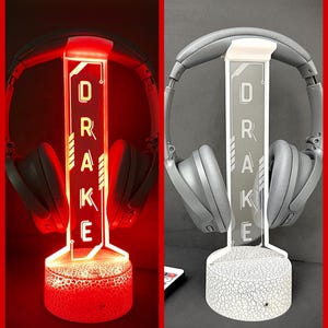 以下が含まれることがあります： 赤色で「DRAKE」の名前が照らされたカスタムヘッドホンスタンド。スタンドはひび割れた地球のベースと長方形のサポートを備えています。グレーのヘッドホンがスタンドに置かれています。スタンドは白い照明でも表示されます。