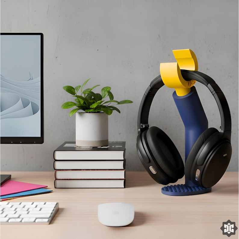 K&ouml;nnte beinhalten: Schwarzer Over-Ear-Kopfh&ouml;rer auf einem blau-gelben Kopfh&ouml;rerst&auml;nder. Eine kleine Topfpflanze steht auf einem Stapel B&uuml;cher. Ein Computermonitor, eine Tastatur und eine Maus sind ebenfalls zu sehen.