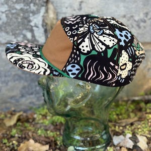 May include: A flat-brimmed baseball cap with a brown bill and a green trim. The cap features a black background with a colorful floral pattern in white, green, and blue. The cap is displayed on a glass head.