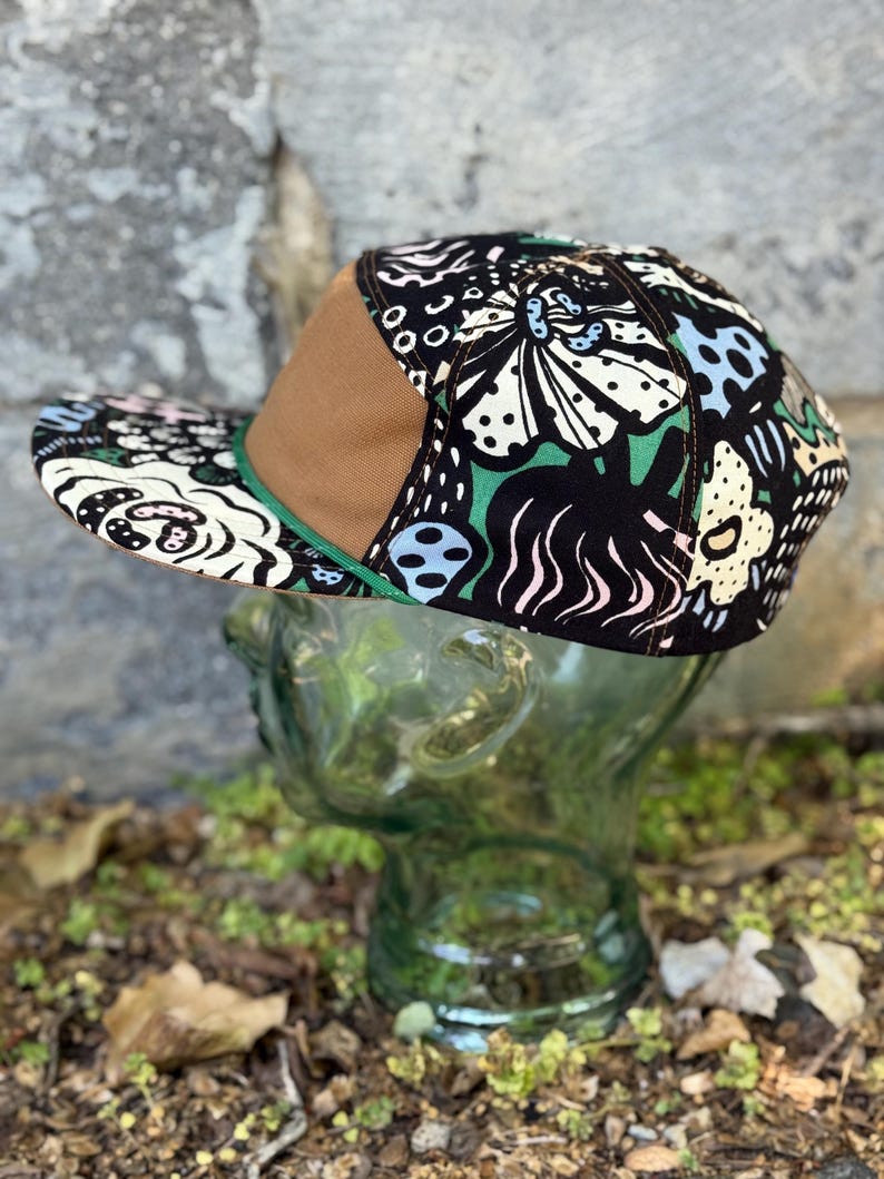 May include: A flat-brimmed baseball cap with a brown bill and a green trim. The cap features a black background with a colorful floral pattern in white, green, and blue. The cap is displayed on a glass head.