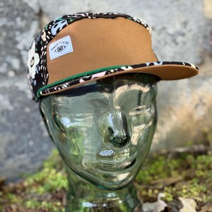 May include: A brown and floral print five-panel cap with a flat brim. The cap features a brown front panel, a black and floral patterned side panel, and a green trim. A white label with text and an eye graphic is on the side.