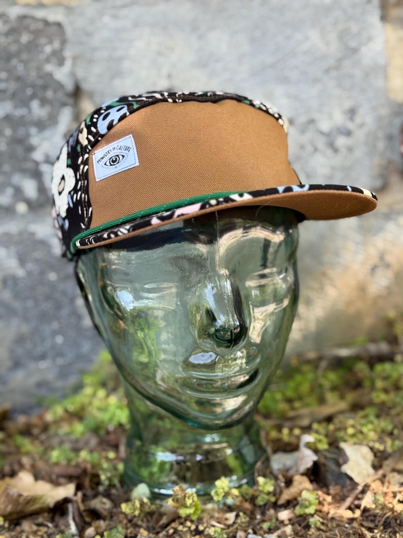 May include: A brown and floral print five-panel cap with a flat brim. The cap features a brown front panel, a black and floral patterned side panel, and a green trim. A white label with text and an eye graphic is on the side.