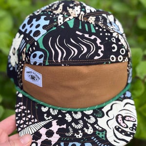 May include: A five-panel cap with a brown bill and a black crown patterned with white, blue, pink, and green abstract designs. A green rope trim accents the bill. A small white tag with text is sewn on the front.