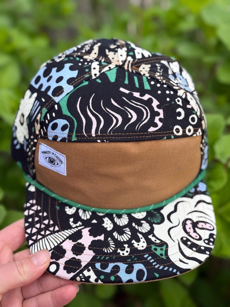 May include: A five-panel cap with a brown bill and a black crown patterned with white, blue, pink, and green abstract designs. A green rope trim accents the bill. A small white tag with text is sewn on the front.
