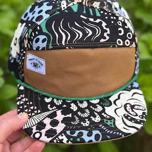 May include: A patterned five-panel cap with a brown brim and a green rope detail. The cap features a black background with a colorful design, including blue, white, and pink shapes. A small patch with the text "Ministry of Culture" is visible.