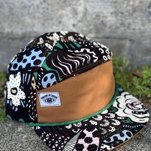 May include: A flat-brimmed cap with a brown front panel and a patterned top featuring black, white, blue, and green designs. The brim is outlined with green trim. A white label reads "Ministry of Culture" with an eye graphic.