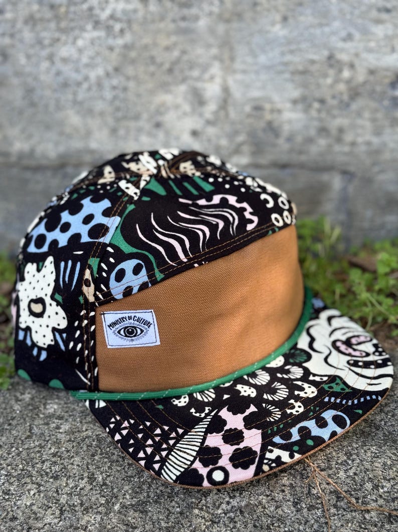 May include: A flat-brimmed cap with a brown front panel and a patterned top featuring black, white, blue, and green designs. The brim is outlined with green trim. A white label reads "Ministry of Culture" with an eye graphic.