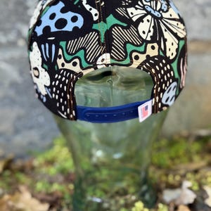 May include: A black snapback hat with a colorful, abstract pattern featuring white, blue, and green shapes. The hat has a blue adjustable strap and a small tag with a flag.