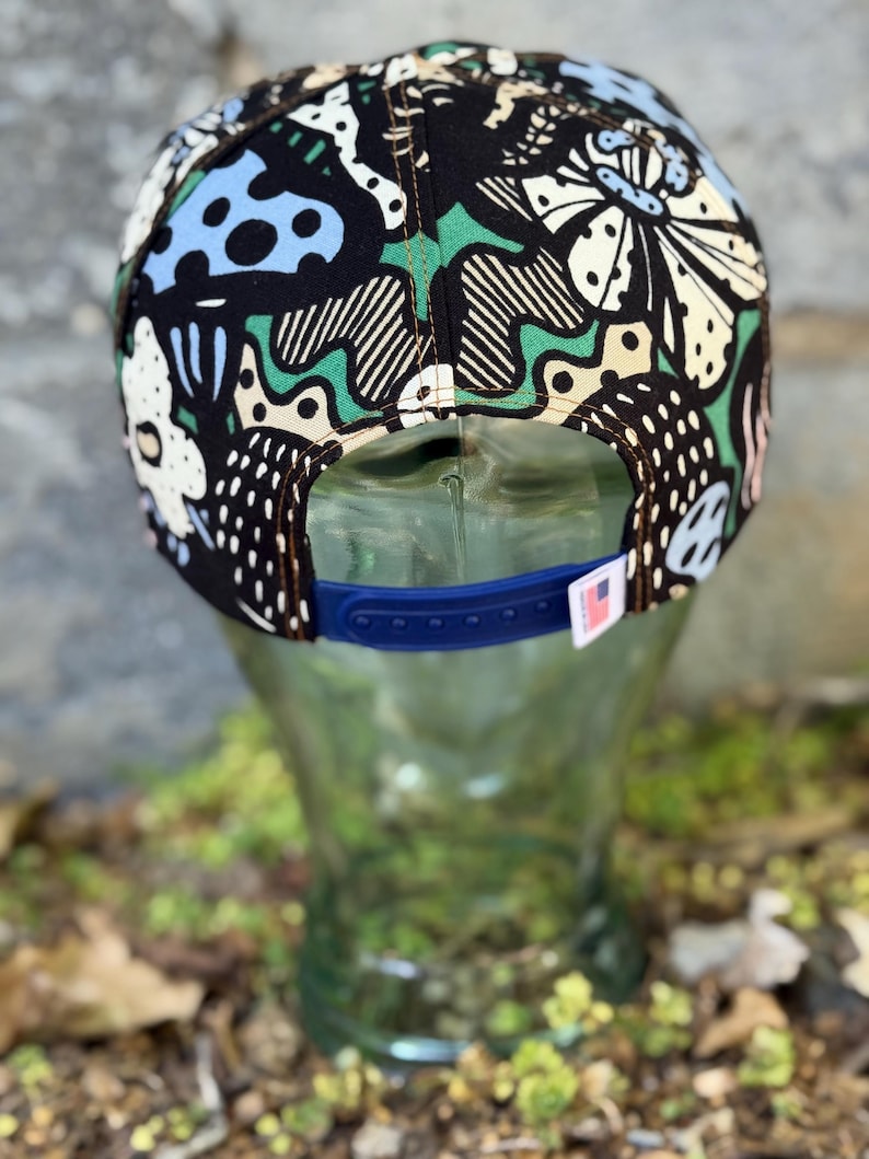May include: A black snapback hat with a colorful, abstract pattern featuring white, blue, and green shapes. The hat has a blue adjustable strap and a small tag with a flag.