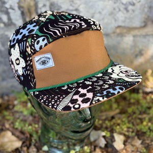 May include: A flat-brimmed cap with a brown front panel and a patterned top featuring black, white, blue, and pink designs. The cap has a green trim and a small label that reads "Ministry of Culture."