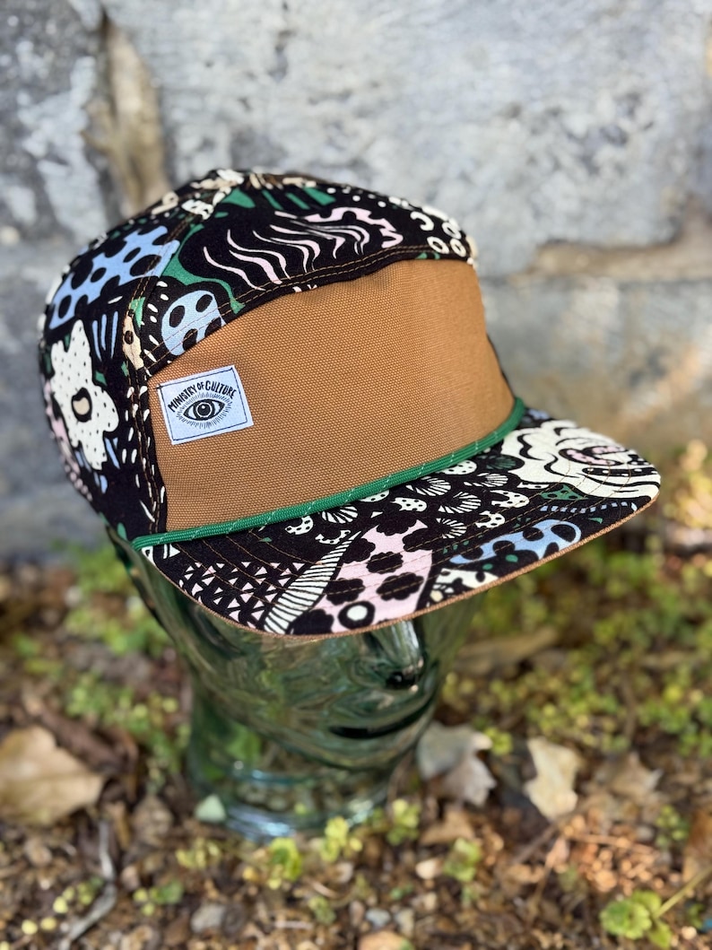 May include: A flat-brimmed cap with a brown front panel and a patterned top featuring black, white, blue, and pink designs. The cap has a green trim and a small label that reads "Ministry of Culture."