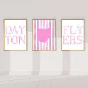 May include: Three framed prints with gold-colored frames. The left print says "DAYTON" in pink striped letters. The center print has a pink Ohio state outline on a pink and white striped background. The right print says "FLYERS" in pink striped letters.