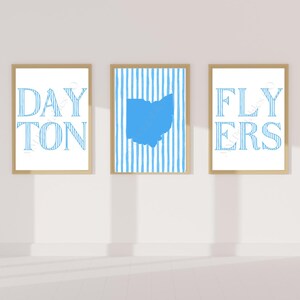 May include: Three framed prints with a light wood finish. The left print says "DAYTON" in blue, the center print has a blue Ohio outline on a striped background, and the right print says "FLYERS" in blue.
